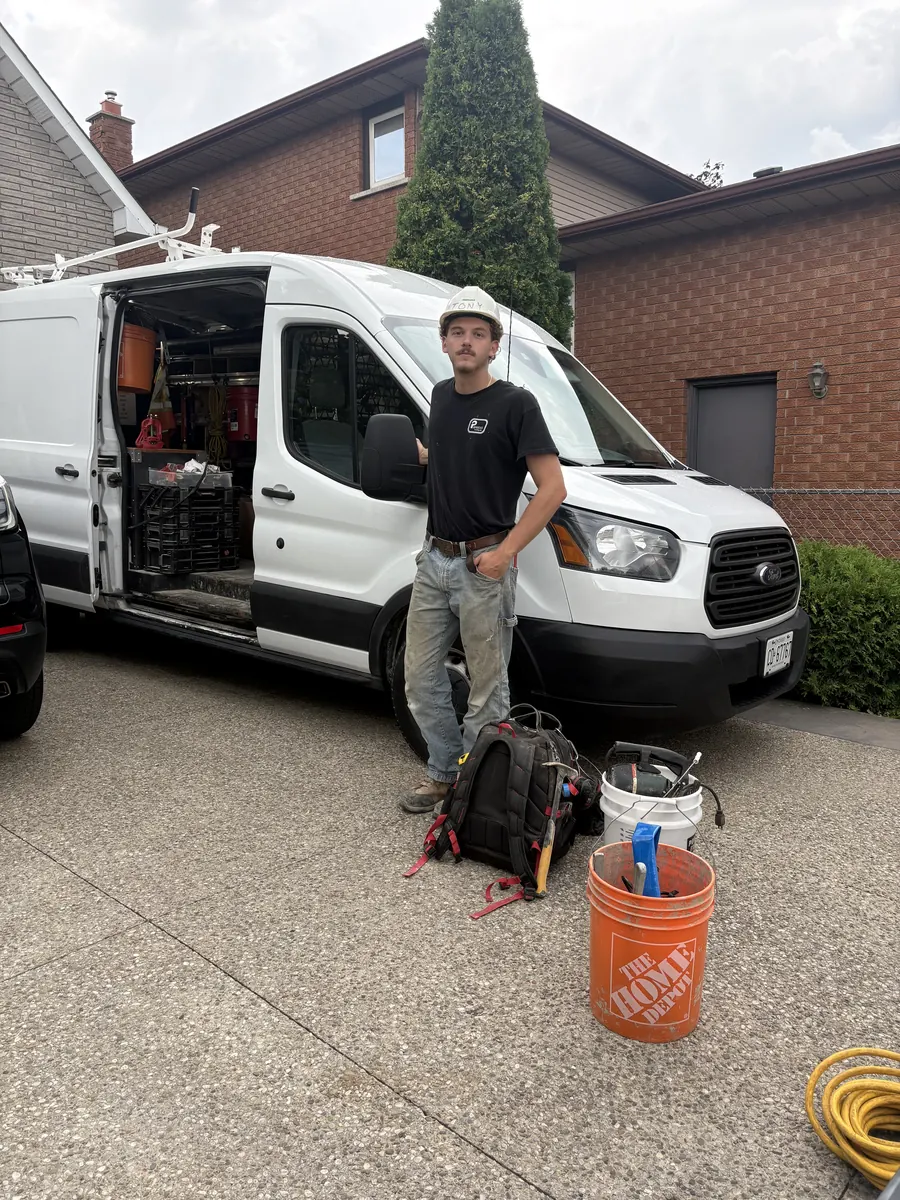 Anthony Panacci, owner of Panacci Plumbing, standing in a driveway beside his white work van with his tool bag and buckets