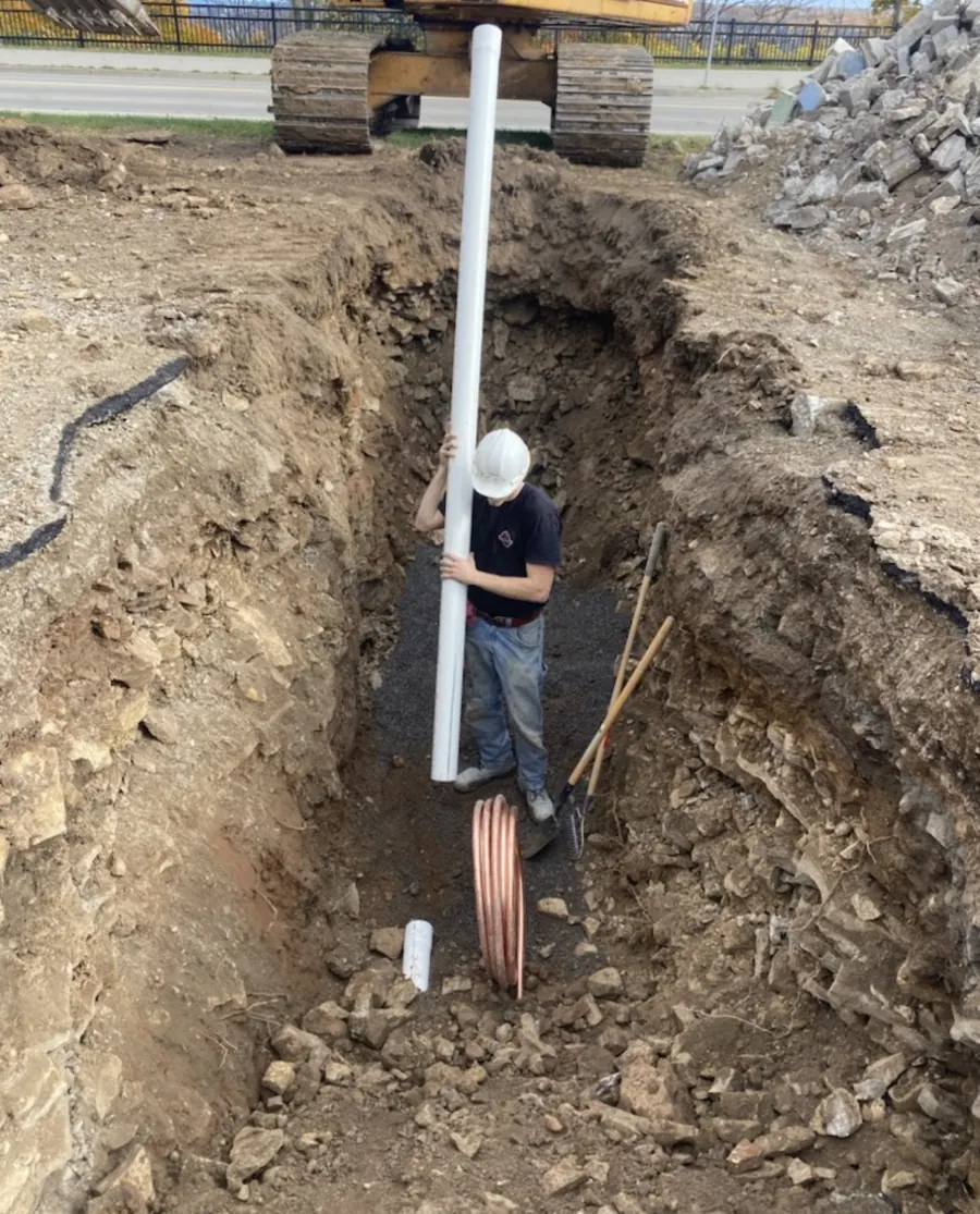 Plumber in an excavated trench setting new underground pipe with a coil of copper water line