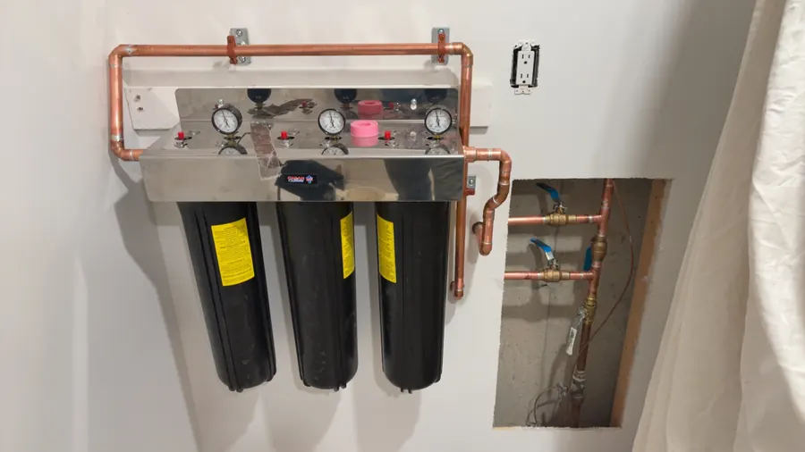 Three-stage whole-home water filtration system with pressure gauges and copper piping mounted on a basement wall